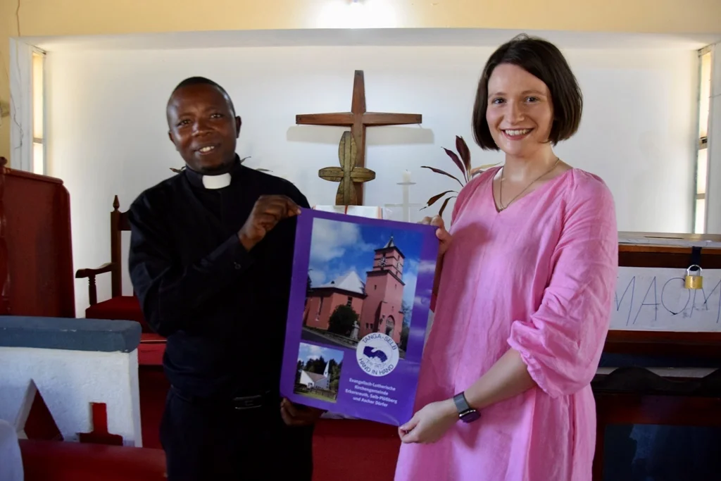 Bild: Anne-Sophie Göbel übergibt das Plakat der Kirchengemeinde Erkersreuth an Pfarrer Tilmilai Shenkalwa der Gemeinde Pangani 