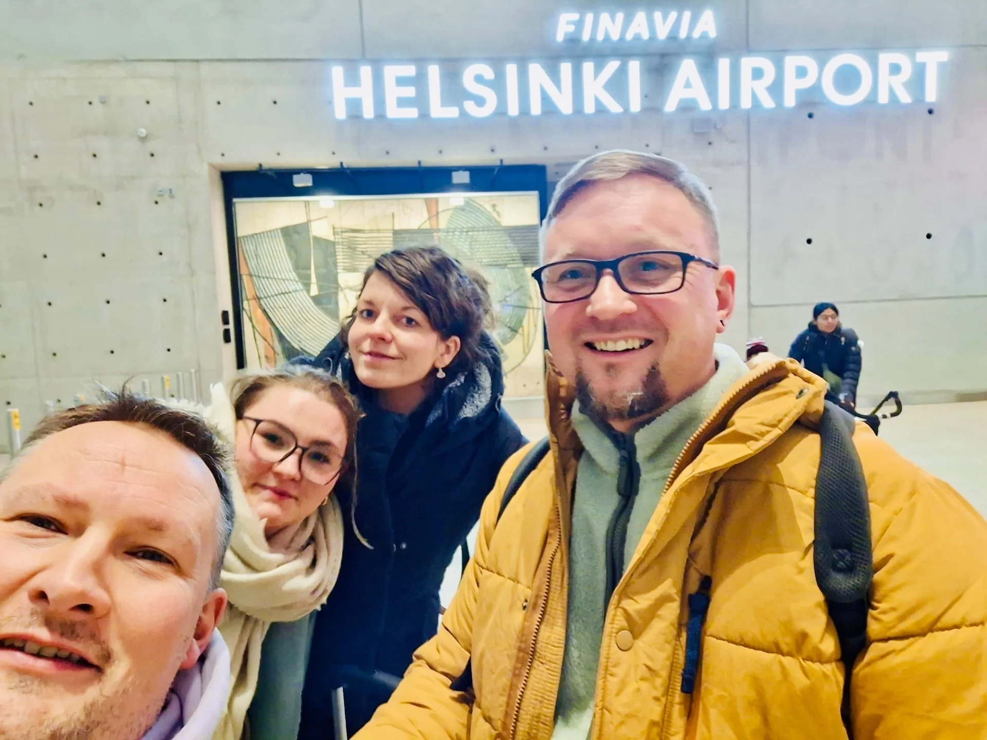 Bild: Vier Personen lächeln, im Hintergrund Schild Flughafen Helsinki