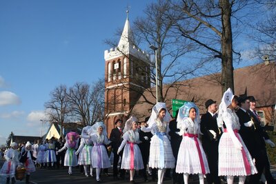 Die Frauen und Mädchen in ihren wunderschönen Tanztrachten und die Männer mit Anzug ziehen beim Fastnachtsumzug durch das Dorf. Im Bild gehen sie an der Kirche vorbei.