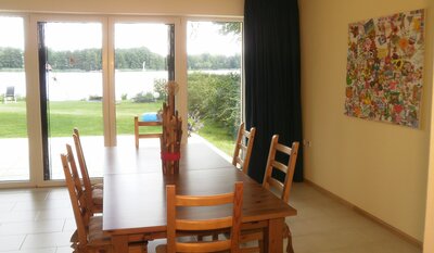 Vorschaubild: Neben der lederbezogenen Sitzgruppe mit Couchtisch steht ein großer Esstisch im kombinierten Schlaf-, Wohn- und Esszimmer mit Ausblick auf den See.