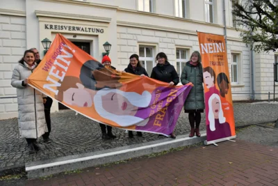 Foto: Bernd Atzenroth/Landkreis Prignitz | Die Fahne wird fürs Hissen vorbereitet. Die Gleichstellungsbeauftragte des Landkreises Prignitz, Christina Rätke (l.), und ihr Pendant in der Stadt Perleberg, Regine Tausendfreund (2.v.r.), hatten sich dafür vor der Kreisverwaltung verabredet.