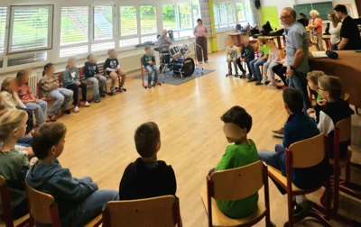 Kinder sitzen in einem großen Kreis im Klassenzimmer, während Erwachsene daneben stehen; in der Mitte des Raums ist ein Schlagzeug aufgestellt.