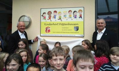 Landrat Bernd Woide und Bürgermeister Manfred Helfrich enthüllen im Beisein der Hortkinder das neue Schild am Eingang zum Kinderhort