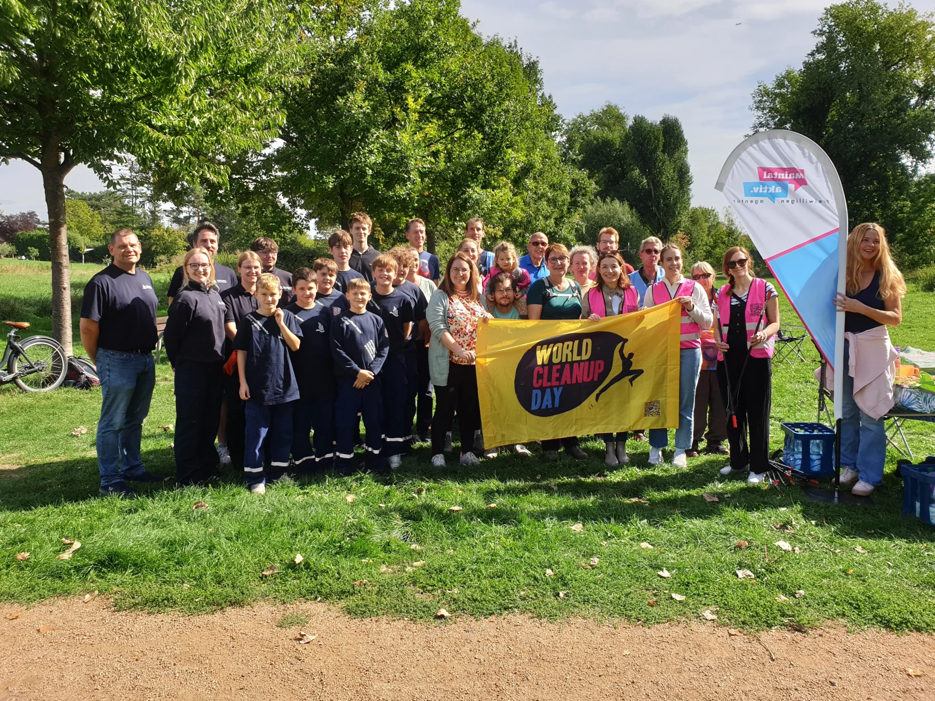 Das Bild zeigt eine Gruppe von Menschen auf einer grüne Wiese. Zwei von ihnen halten ein gelbes Banner hoch, auf dem "World Cleanup Day" steht.
