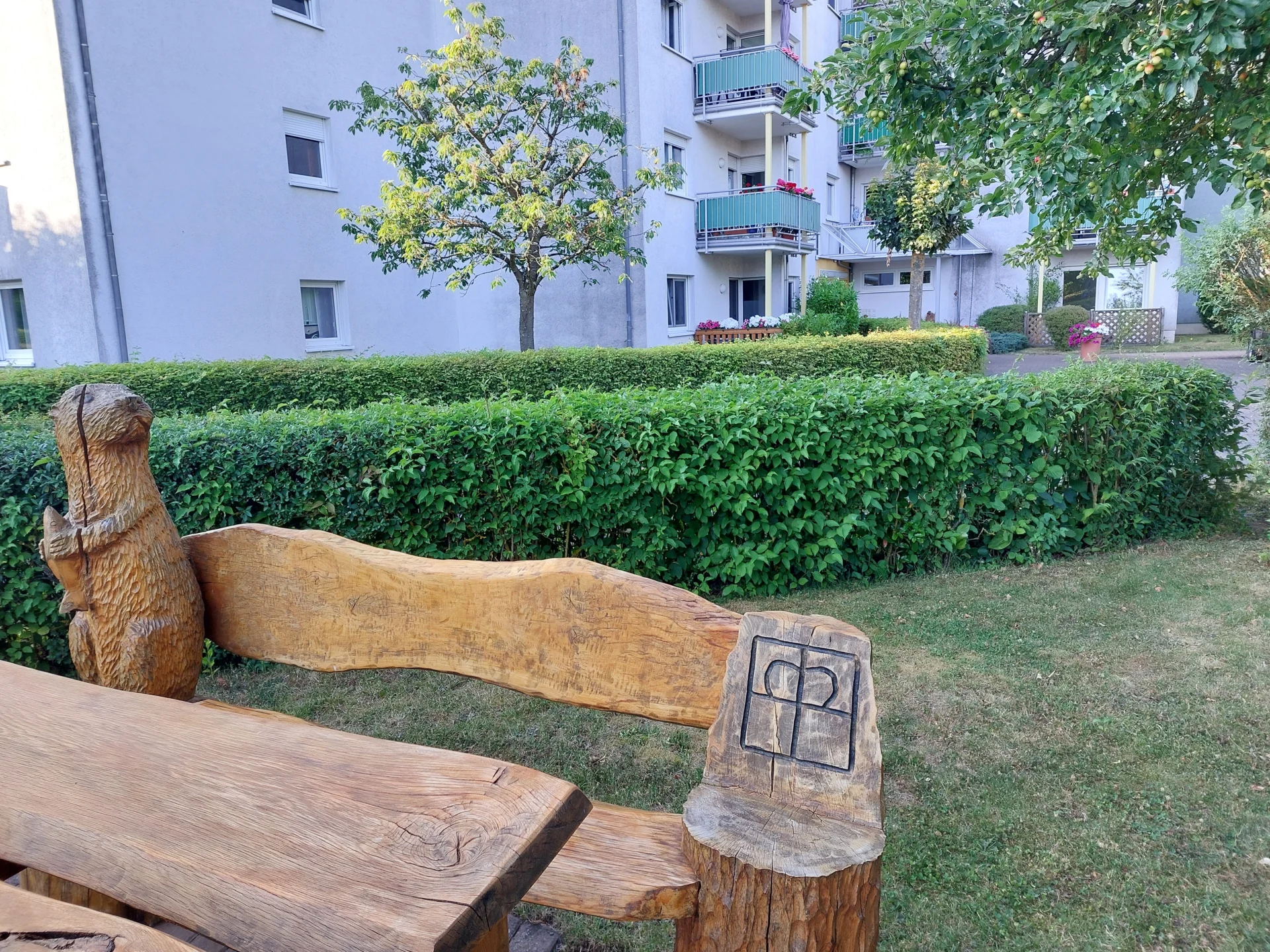 Sitzbank aus Holz, in die das Diakonie-Kreuz eingefräst ist. Im Hintergrund ist die Wohnanlage zu sehen