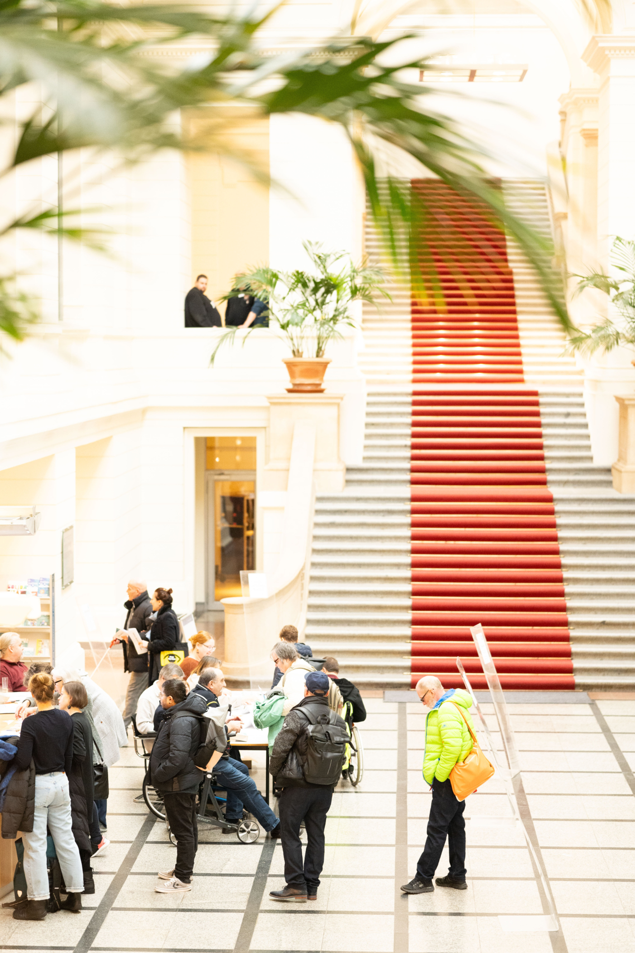 Bild: Erstes Willkommen am Einlass (mehrere Personen warten am Empfang, Bild mit Blick auf die große Treppe mit rotem, mittigem Teppich)