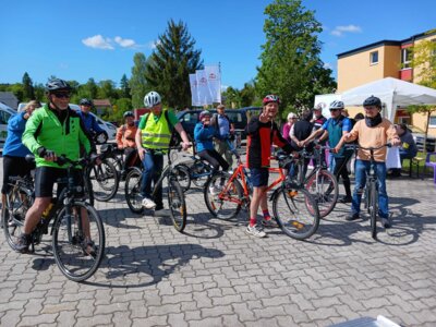 Zehn Menschen sitzen auf speziellen Fahrr&auml;dern f&uuml;r Menschen mit Handicap und schauen in die Kamera, zeigen Peace-Zeichen und l&auml;cheln. Im Hintergrung sind St&auml;nde zu sehen, die auf dem Mobilit&auml;tstag vertreten waren. Das Wetter auf dem Foto ist sonnig.  (Bild vergr&ouml;&szlig;ern)