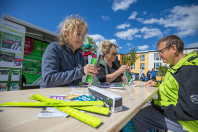 Zwei Frauen vom Verkehrsverbund Vogtland sitezn an einem Tisch. Sie zeigen einem Mann, wie sie einen Bus aus Pappe zusammenbasteln. Im Hintergrund steht ein gr&uuml;ner Bus der Verkehrsbetriebe.  (Bild vergr&ouml;&szlig;ern)