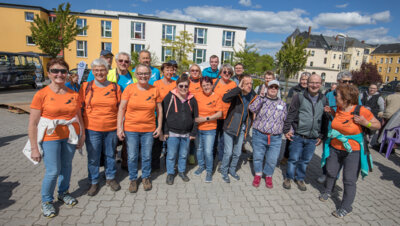 Die Wandergruppe Falkenstein ist mit 21 Personen in orangen T-Shirts zu sehen. Sie stehen im Halbkreis, l&auml;cheln in die Kamera und winken. Es ist sonniges Wetter.  (Bild vergr&ouml;&szlig;ern)