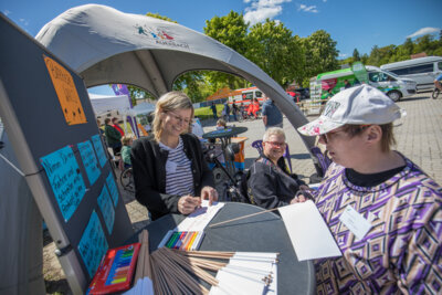 Drei Frauen, davon eine im Rollstuhl, schreiben auf Fahnen und l&auml;cheln in die Kamera. Es sind eine Pinnwand und ein Pavillon der Stadt Auerbach im Hintergrund zu sehen.  (Bild vergr&ouml;&szlig;ern)