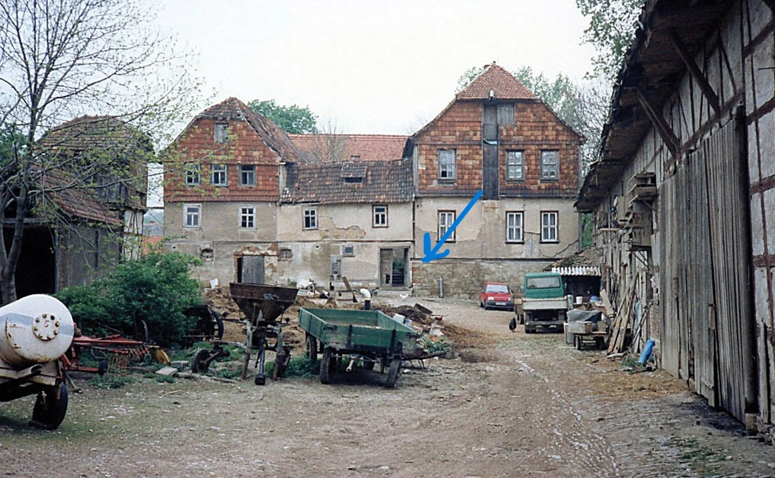 Bild: Ein Foto aus der Zeit vor dem Zusammenbruch der letzten Scheune (rechts im Bild). Die Pfeilspitze zeigt auf die Stelle, an der heute eine Tafel mit Erklärungen angebracht ist.