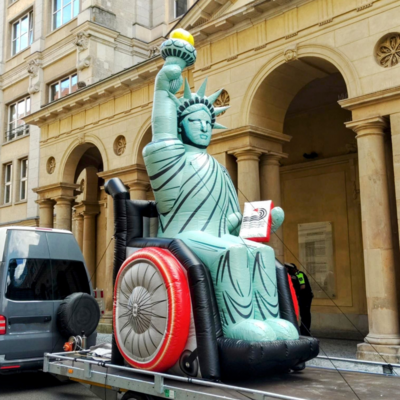 Die Freiheitsstatue mit ihrer Krone, mit ihrer Fackel sitzt in einem Rollstuhl. Das ganze Objekt ist aus Plastik und mit Luft aufgeblasen.