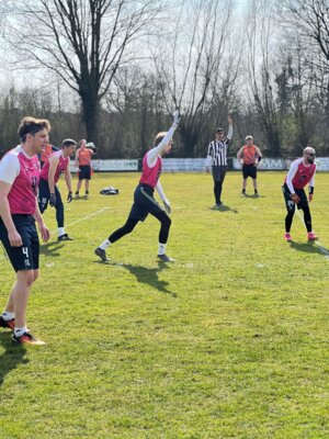Spieler der Waldenau Kraken in pinkfarbenen Trikots stehen in der Verteidigungsformation auf dem Feld. Ein Spieler hebt den Arm zur Signalisierung. Im Hintergrund steht ein Schiedsrichter mit erhobener Hand und gegnerische Spieler in orangefarbenen Trikots bereiten sich auf den Spielzug vor.