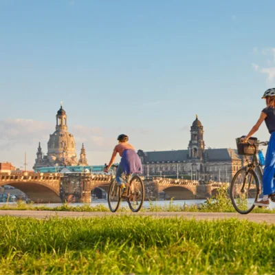 Der Elbradweg mit Blick auf die Brücken und die Frauenkirche, auf dem Weg zwei Fahrradfahrende mit Helmen.