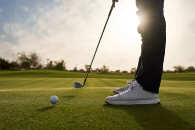 Person steht mit weißen Sportschuhen auf dem Golfplatz und hält Golfschläger neben Golfball auf dem Grün