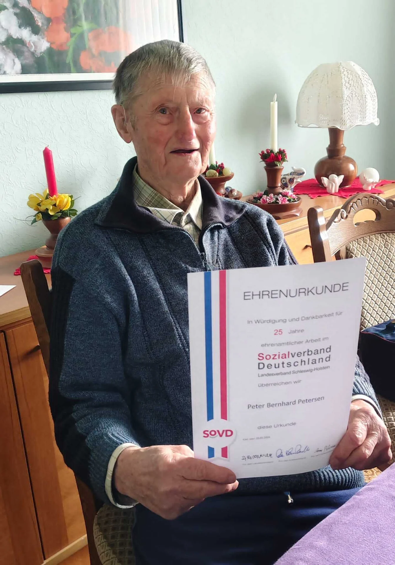 Bild: Peter Bernhard Petersen (92 Jahre) trägt seit über 25 Jahren, treu Monat für Monat, die SoVD-Zeitung bei den Mitgliedern im Ortsteil Rosenkranz aus. Als Dank für diese ehrenamtliche Tätigkeit besuchte ihn die Ortsvorsitzende Anne Petersen zu Hause und überreichte die entsprechende Urkunde, die er stolz auf dem Foto präsentiert, mit Ehrenschild und Präsent.