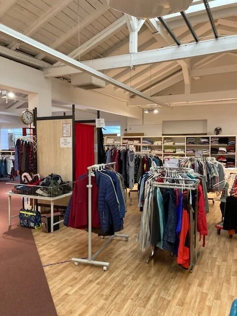 Foto: An Kleiderstangen h&auml;ngen Jacken, Pullover und Hosen. Im Hintergrund sieht man regale, in denen T-Shirts liegen. Man kann eine Umkleidekabine sehen.  (Bild vergr&ouml;&szlig;ern)
