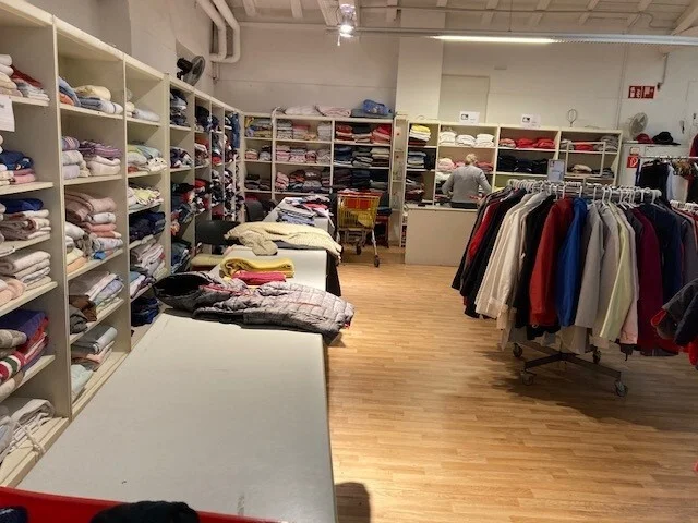 Foto: In Regalen liegen viele Pullover und T-Shirts. An Kleiderst&auml;ndern h&auml;ngen Jacken.  (Bild vergr&ouml;&szlig;ern)