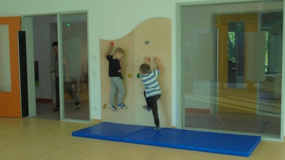Bild: Der Sportraum ist hell und groß und bietet Vieles, was zur sportlichen Aktivität anregt. Eine Kletterwand für die Hand-Fuß-Koordination, Leitern, Koppelelemente und einen Kasten, sowie Matten, Bälle, Reifen, Bänder und verschiedene Treppenelemente dienen den Kindern dazu sich sportlich zu betätigen. Gemeinsame Bewegungsspiele und Bewegung nach Musik fördern das Takt-und Rhythmusgefühl. Die Freude und der Spaß an körperlicher Betätigung soll geweckt werden.