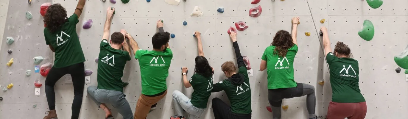 Sieben Personen mit inklusiv aktiv T-Shirts hängen an der Kletterwand