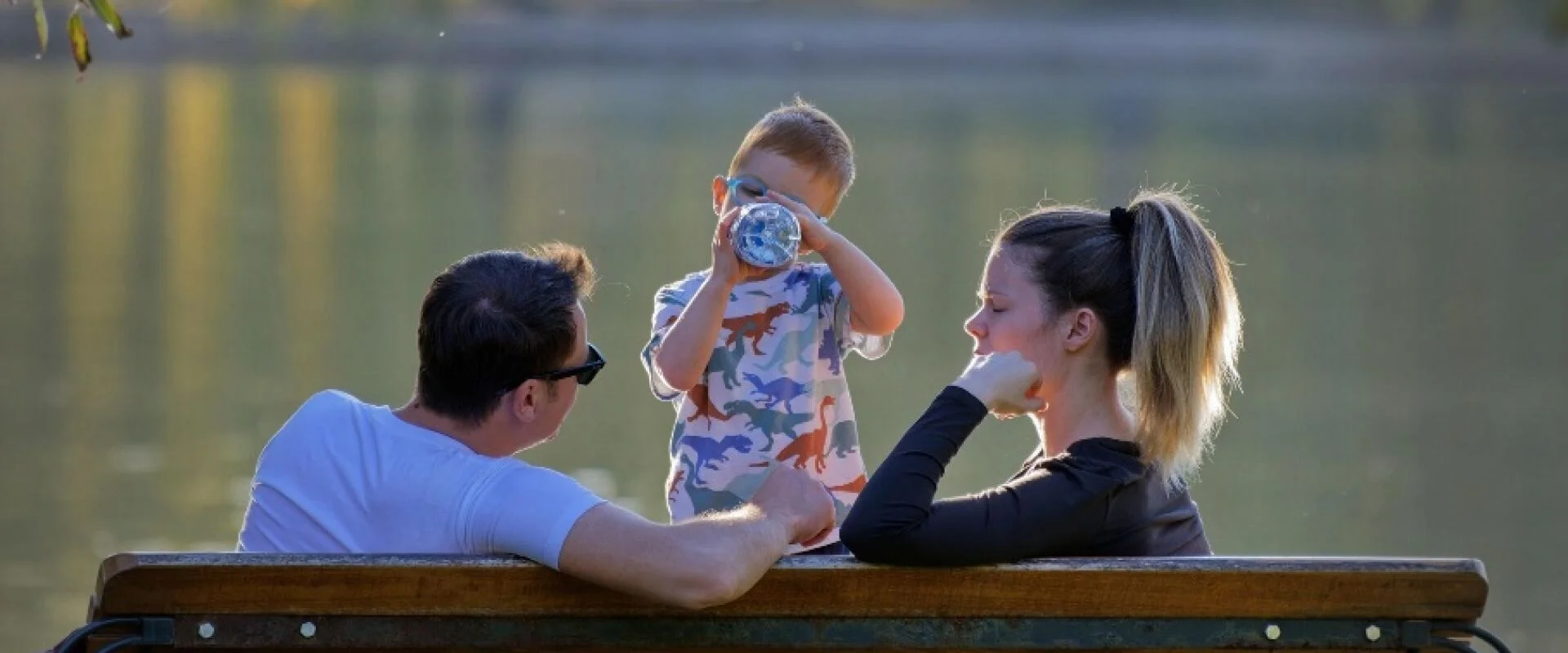 Foto: Mama, Papa und Kind sitzen auf einer Bank am See. Das Kind trinkt aus einer Flasche.
