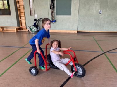 Zwei Schüler:innen fahren auf einem neu gekauften Dreirad durch die Turnhalle