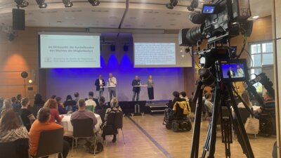 Foto aus dem Veranstaltungsraum. Gezeigt ist Henning Knapheide mit 3 weiteren Personen im Interview auf der Bühne.  Oberhalb der Bühne befinden sich Bildschirme mit Gebärden- und Schriftdolmetschung. Vor der Bühne sind viele Zuschauerinnen und Zuschauer zu sehen und eine Kamera ist auf die Bühne gerichtet.