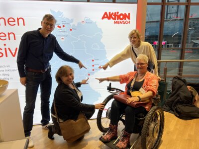 Ein Mann, zwei der Korrdinatorinnen und eine Frau im Rollstuhl stehen vor einer Leinwand und zweigen mit ihren Zeigefinger auf eine projezierte Karte. Es ist die Deutschlandkarte und sie zeigen auf die Kommunen in Sachsen, die am Projekt "Inklusion vor Ort" teilnehmen. (Bild vergr&ouml;&szlig;ern)