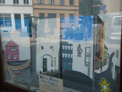Foto: Rolandstadt Perleberg/Renè Hill | Während sich im Schaufenster das Leben auf dem Schuhmarkt widerspiegelt, sind im Schaufenster die künstlerischen Arbeiten der Oberschülerinnen zu sehen.