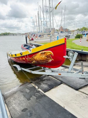 Der ZK10- Kutter Barther Greif (das Boot hat einen roten Grundanstrich, darauf das Profil eines Greifs als Grafitti, oben am Boot ein gelber Streifen), auf dem Trailer in der Slippanlage, das Boot ist mitv dem Heck bereits im Wasser (Bild vergr&ouml;&szlig;ern)