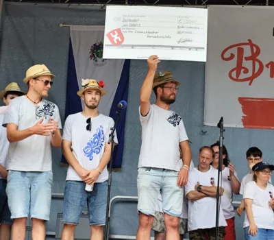 Foto: Auf dem Foto sieht man im Vordergrund drei „Liewerschbescher Kerweborschde“. Einer hält einen großen Spendencheck hoch. Im Hintergrund links ist ein Teil der D!-Gemeinschaft zu sehen. Foto zur Meldung: 500 Euro Spende der Liewerschbescher Kerweborschde