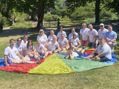 Foto: Auf dem Foto sieht man die Mitglieder der D!-Gemeinschaft, die am Samstag beim Picknick dabei waren. Alle sitzen auf einem großen, regenbogenfarbenen Schwungtuch. Foto zur Meldung: Ein Wochenende voller Gemeinschaft und Musik