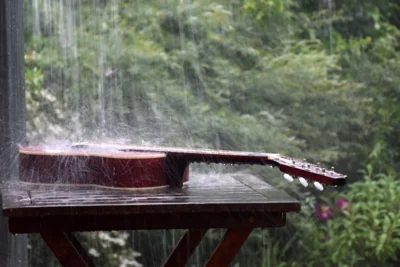 Eine rote Gitarre liegt auf einem Holztisch im Garten, und starker Regen prasselt auf sie herab.