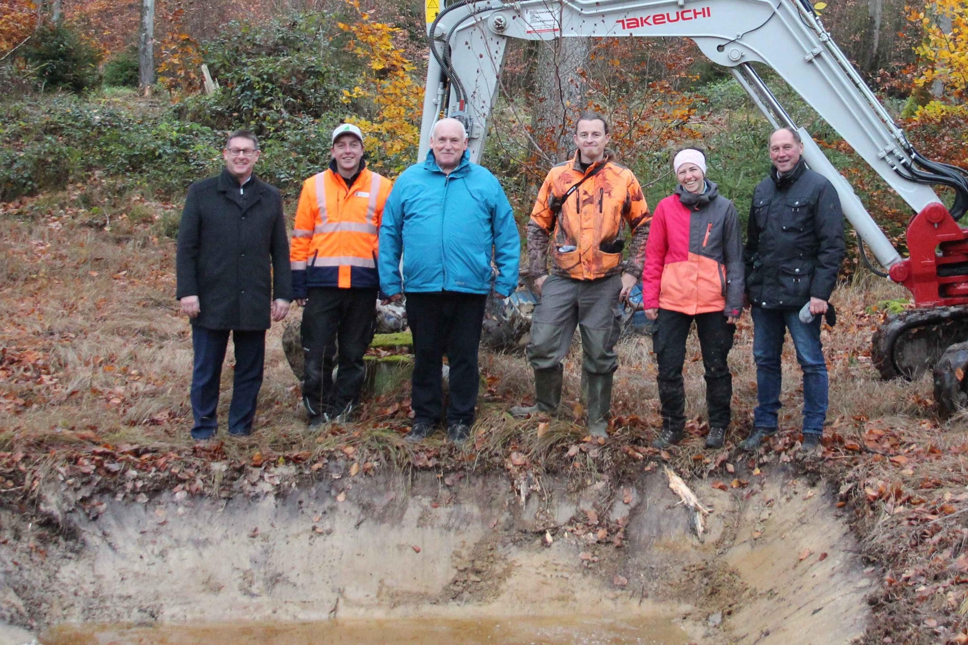 Sechs Personen in Outdoor-Kleidung stehen nebeneinander vor herbstlichem Wald und Baumaschine