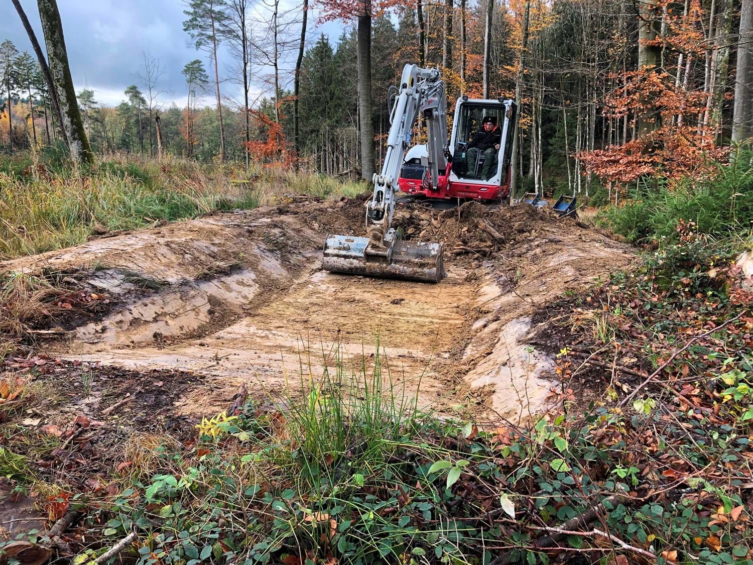 Bagger mit ausgefahrenem Arm arbeitet auf einem freigelegten Erdweg im Wald mit herbstlich gefärbtem Laub.