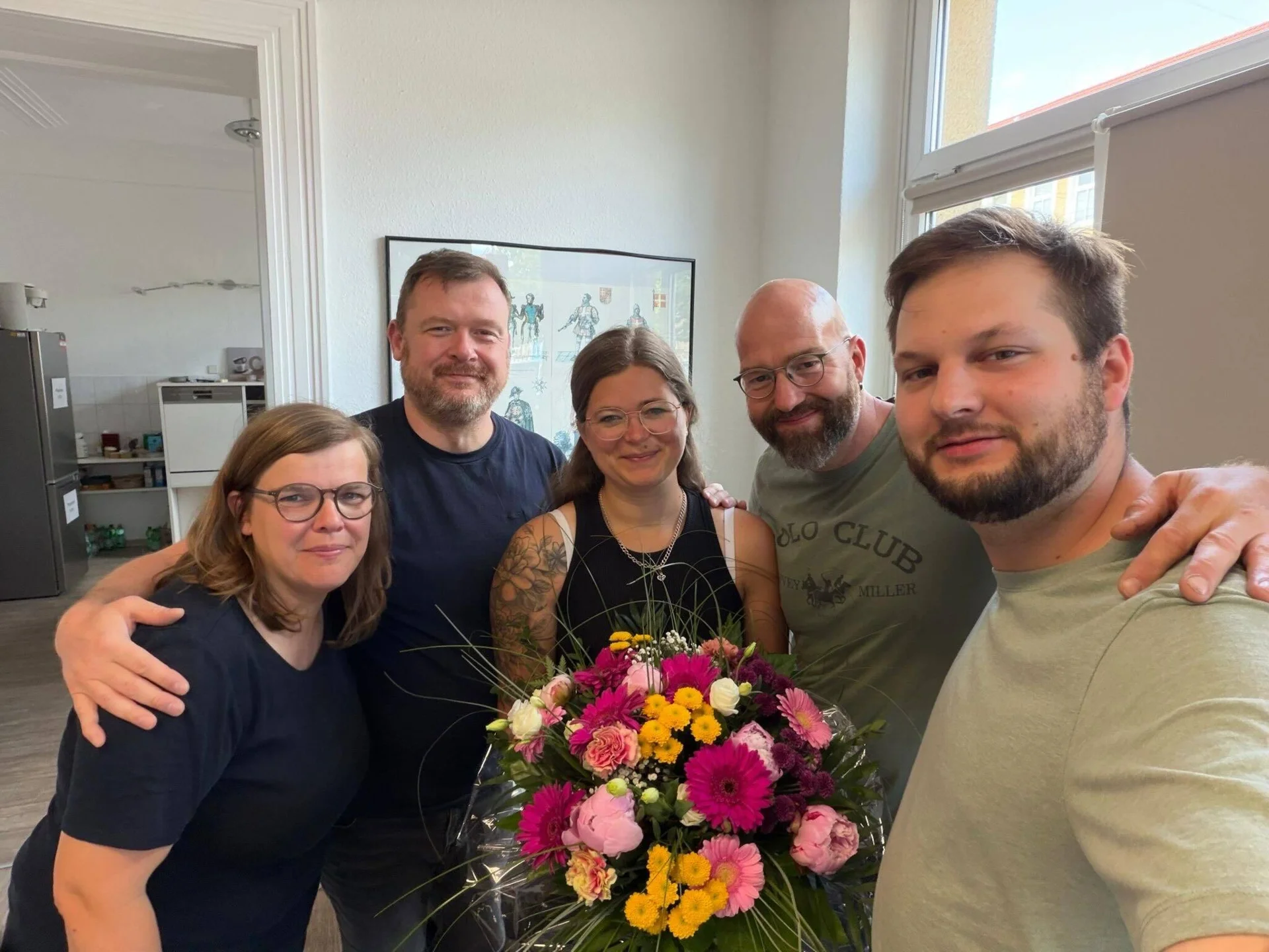 Gruppenfoto mit der neuen Projektkoordinatorin. Fünf Personen stehen gemeinsam in einem Büro und halten einen bunten Blumenstrauß zur Begrüßung.