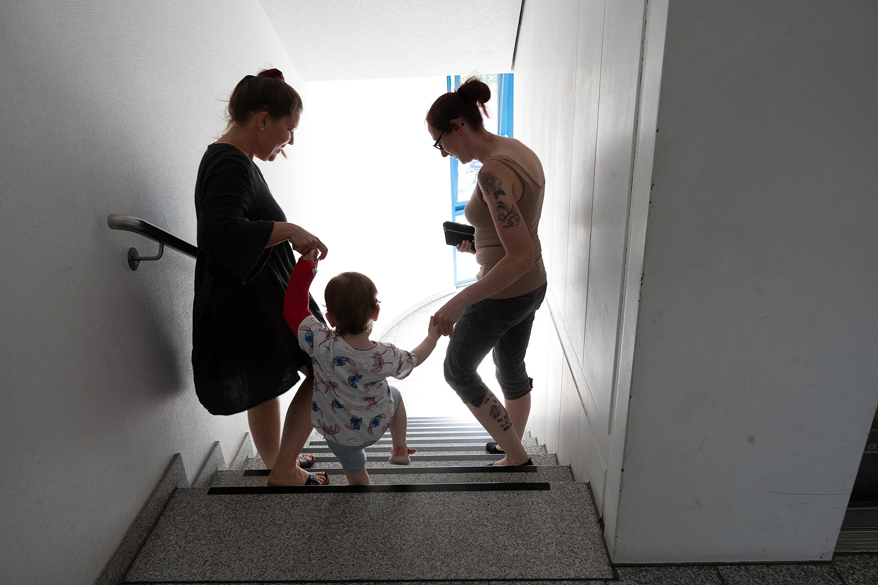 Zwei Frauen gehen gemeinsam mit einem Kleinkind eine Treppe hinunter. Jede von ihnen hält das Kind an einer Hand und schaut auf das KInd.
