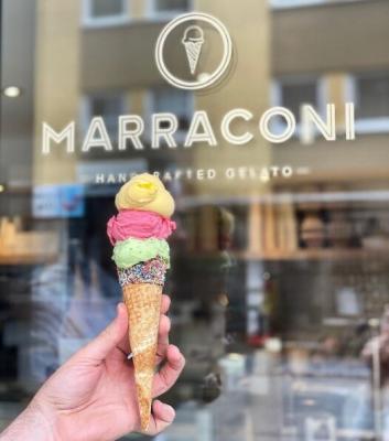 Auf dem Foto sieht man eine Hand, die eine Eistüte mit je einer gelben, einer roten und einer grünen Kugel Eis hält. Dahinter ist auf dem Schaufenster der Schriftzug Marraconi zu lesen.