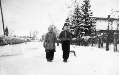 Auf der zugeschneiten Backemoorer Straße laufen um 1958 Hermine Erfeling (links) und Elise Weber mit einem Schlitten. Rechts im Hintergrund sieht man das Haus der Familie Groenefeld. Ganz hinten links sieht man ein Haus, in dem Habbo und Altjeline Fröhling ein Lebensmittelgeschäft mit Bäckerei betrieb. (Bildquelle: Hermine Oltmanns/Repro Groeneveld). (Bild vergrößern)