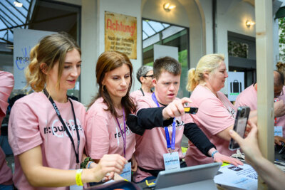 Vier Personen stehen nebeneinander. Sie tragen pinke T-Shirts mit der Aufschrift "I care". Eine Person scannt ein Ticket.. (Bild vergrößern)