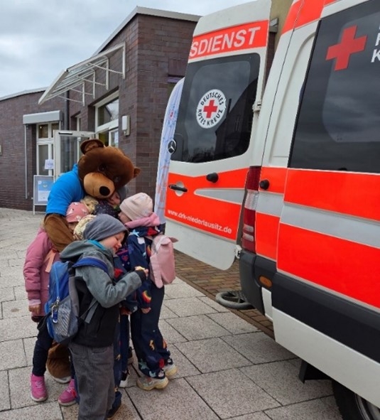Großer Teddybär und Kitakinder vor Krankenwagen