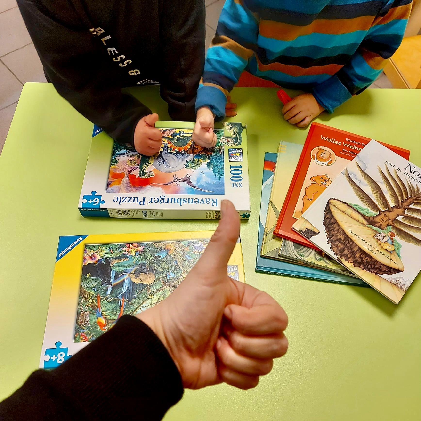 Auf dem Foto sieht man vier Bücher und zwei Puzzles auf einem Tisch liegen, sowie drei Arme von Kindern, die drei hochgestreckten Daumen über die Materialien halten.