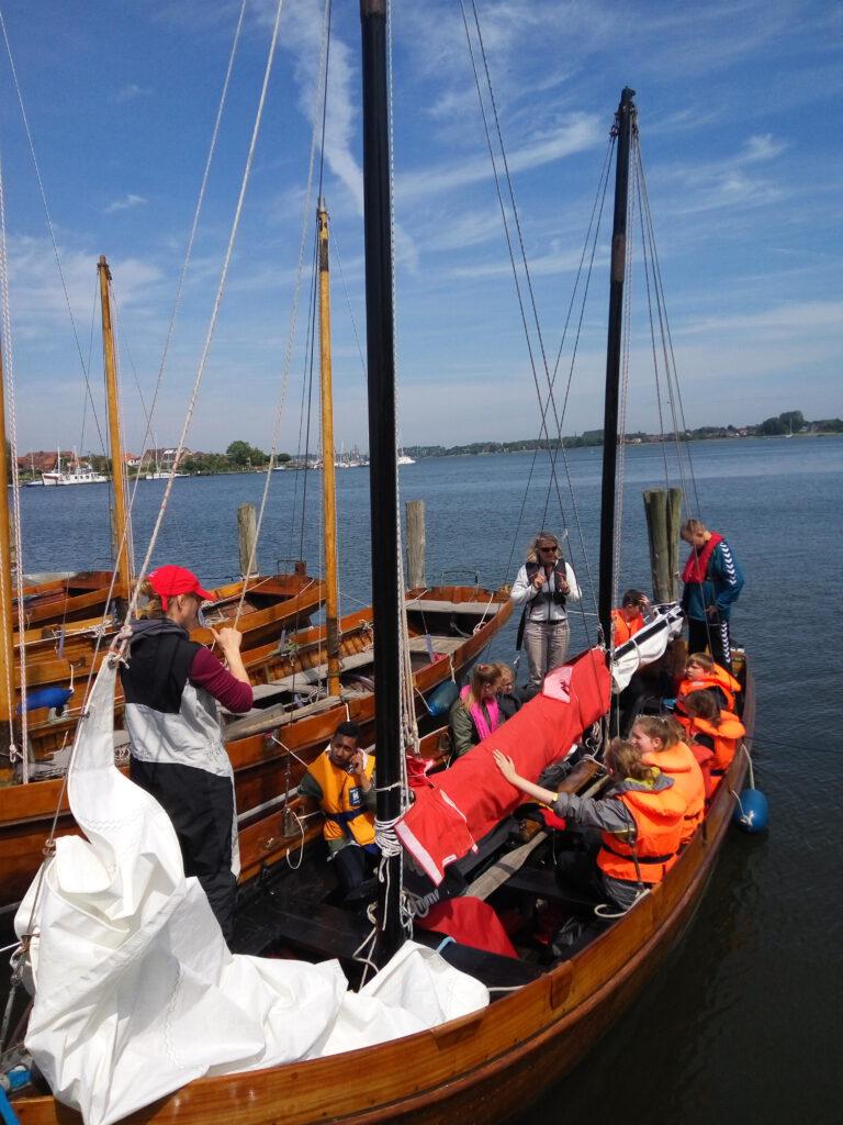 Man sieht ein Segelboot aus Holz, in dem Kinder links und rechts an der Sitzen. Die Kinder tragen orangene Schwimmwesten. Zwischen den Kindern befindet sich ein herabgelassenes, weißes Segel, das zur Hälfte von einer roten Schutzhülle abgedeckt ist. Am Bug des Bootes steht eine Person, die den Kindern etwas erklärt. Hinter dieser Person befindet sich ein weiteres herabgelassenes, weißes Segel. Am Heck des Bootes stehen zwei weitere Personen.   Links neben dem Boot sieht man drei weitere Boote. Diese sind leer. Im Hintergrund des Bildes ist die Schlei zu sehen.