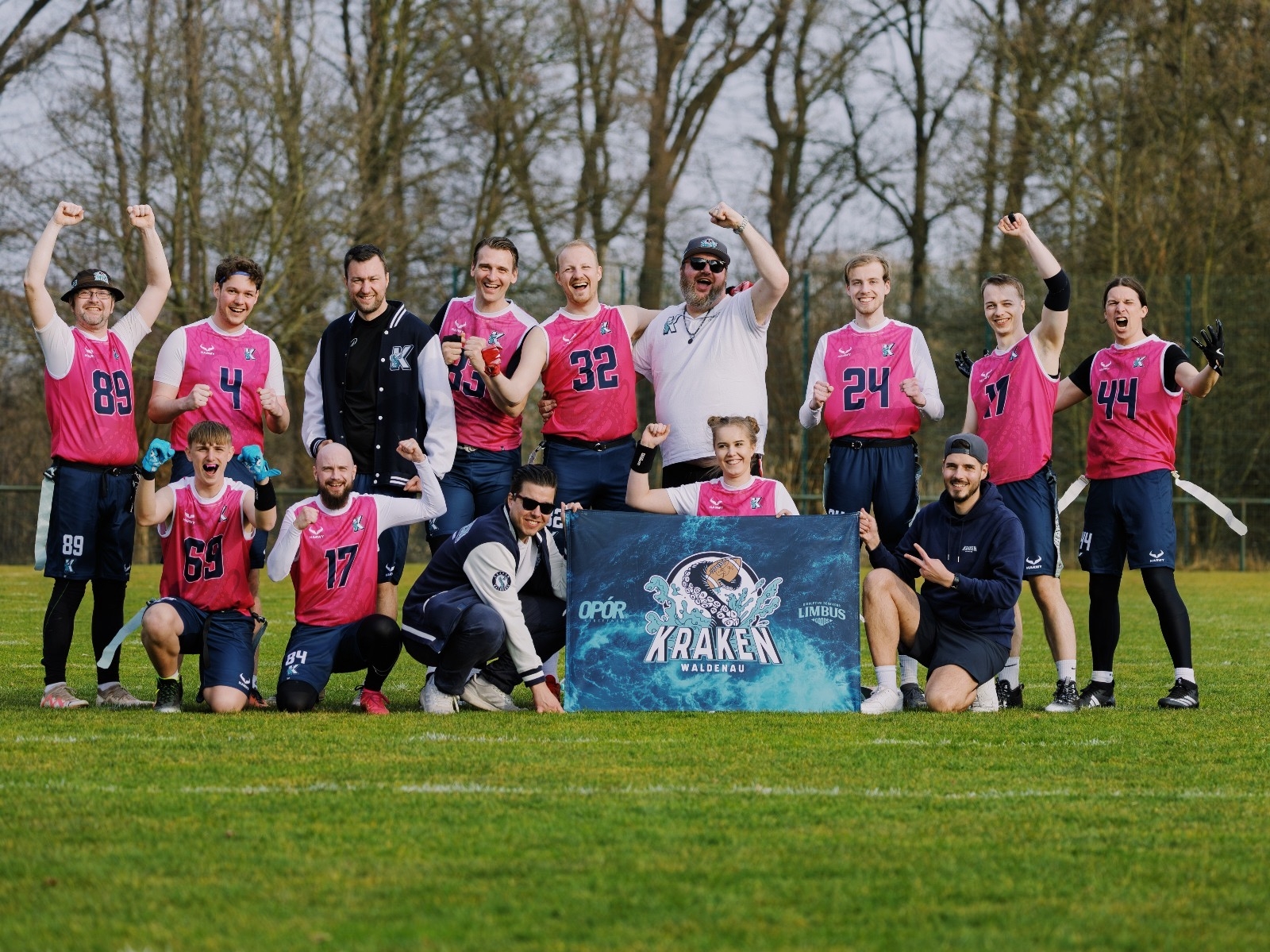 Das Team der Waldenau Kraken posiert jubelnd auf einem Sportplatz. Alle tragen pinkfarbene Flag Football Trikots mit blauen Shorts und lächeln oder zeigen jubelnde Gesten in die Kamera. Vor der Gruppe halten zwei Personen ein Banner mit dem Logo der Waldenau Kraken, das einen stilisierten Krakenkopf zeigt. Im Hintergrund sind Bäume ohne Blätter zu sehen, es scheint ein klarer Tag zu sein.