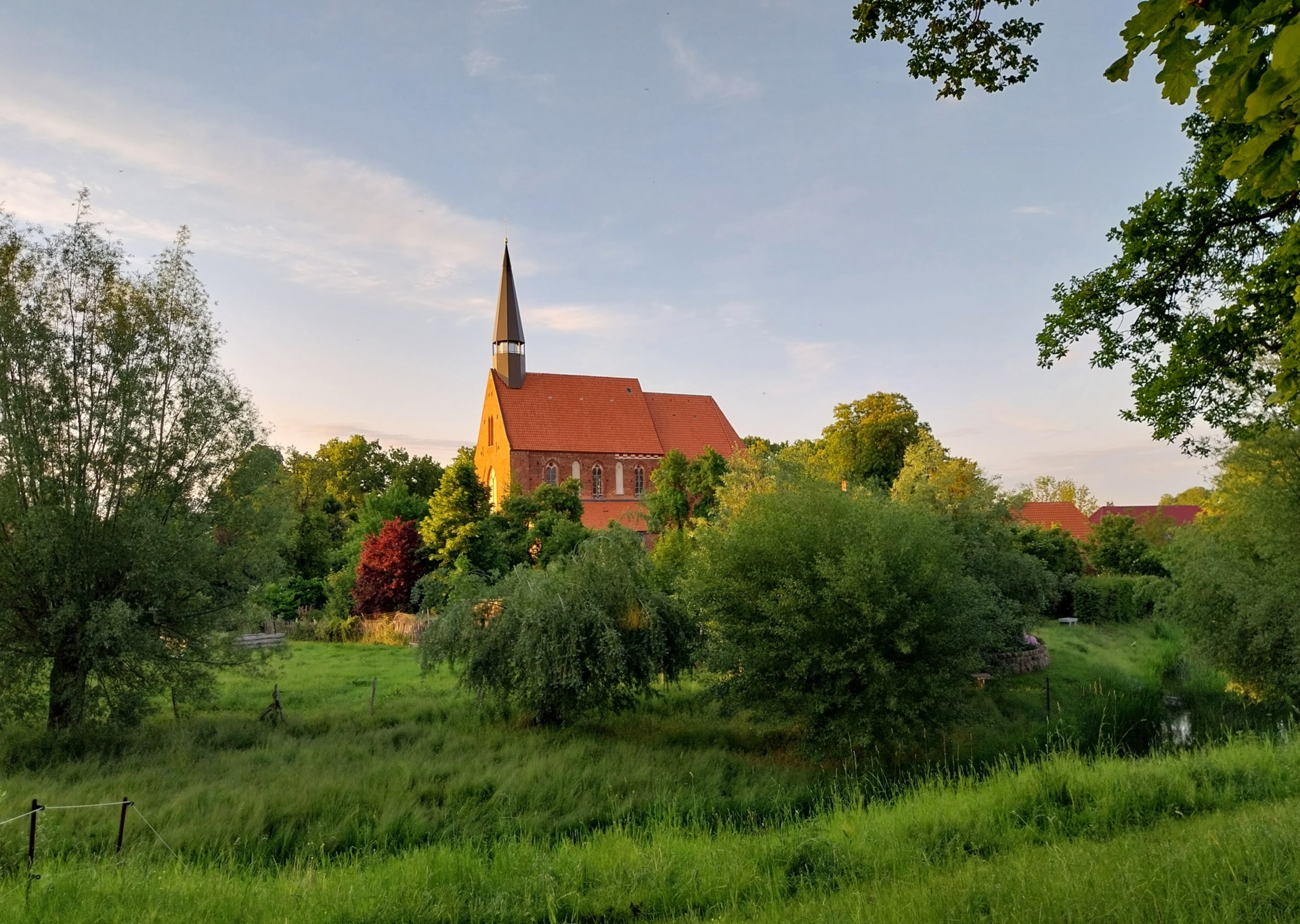 Das Bild zeigt die Starkower Backsteinkirche mit einem hohen, dunklen Turm und einem roten Ziegeldach. Die Kirche steht inmitten einer grünen Landschaft mit hohen Bäumen und niedrigem Gras. Das Licht scheint von der Seite zu kommen, was die Kirche und die umliegenden Bäume beleuchtet. Der Himmel ist leicht bewölkt. Das Gesamtbild wirkt ruhig und idyllisch, fast wie eine Postkartenansicht in der ländlichen Gegend von Starkow.