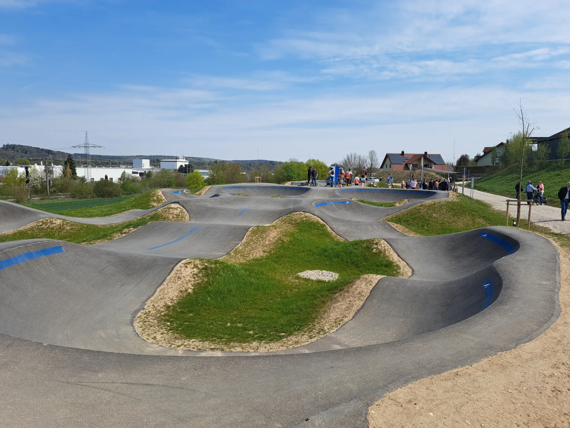 Pumptrack mit geschwungenen Asphaltstrecken und grasbewachsenen Hügeln unter blauem Himmel
