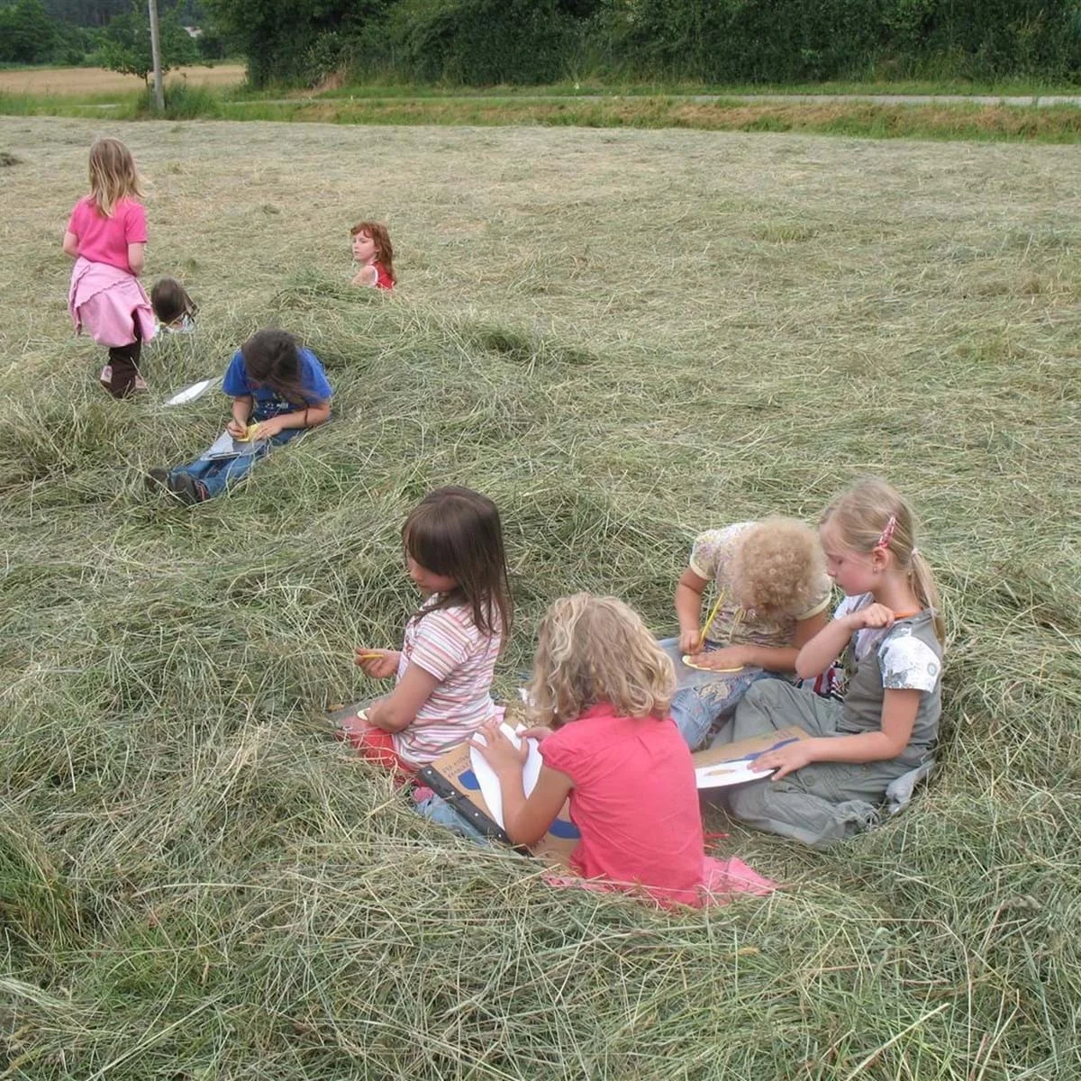 Mehrere Kinder sitzen und stehen auf einem Feld mit hohem Gras, einige halten Schreibtafeln oder Bücher