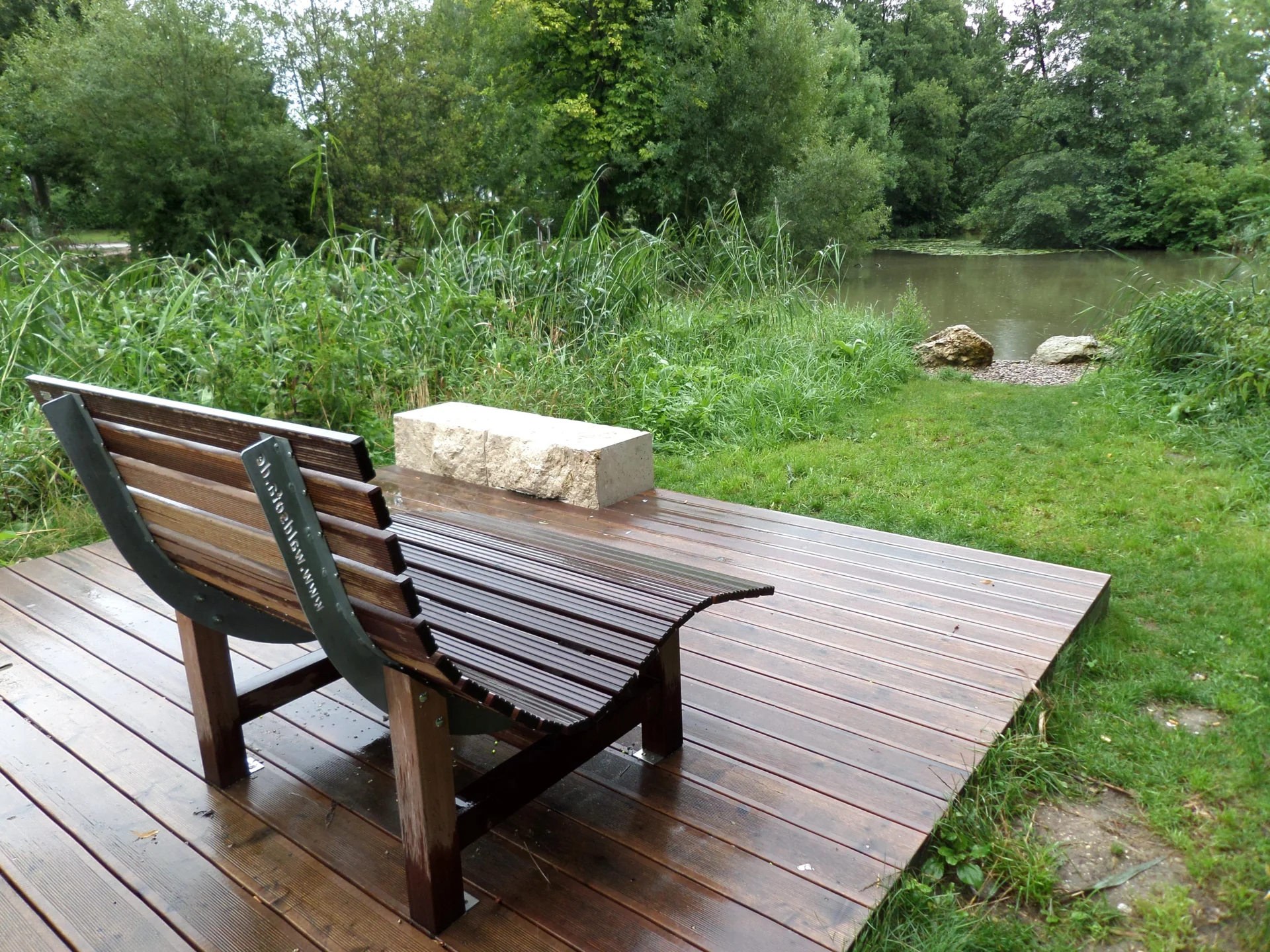 Holzbank mit gebogenem Sitz auf Holzplattform am Ufer eines Teichs mit Gras und Büschen im Hintergrund