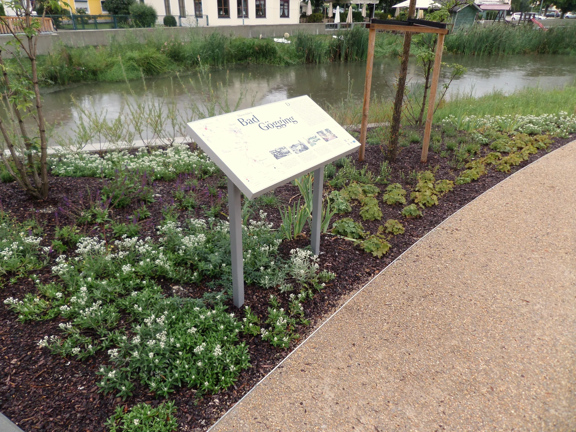 Infotafel mit der Überschrift „Bad Gögging“ in einem bepflanzten Beet neben einem Weg und einem Fluss