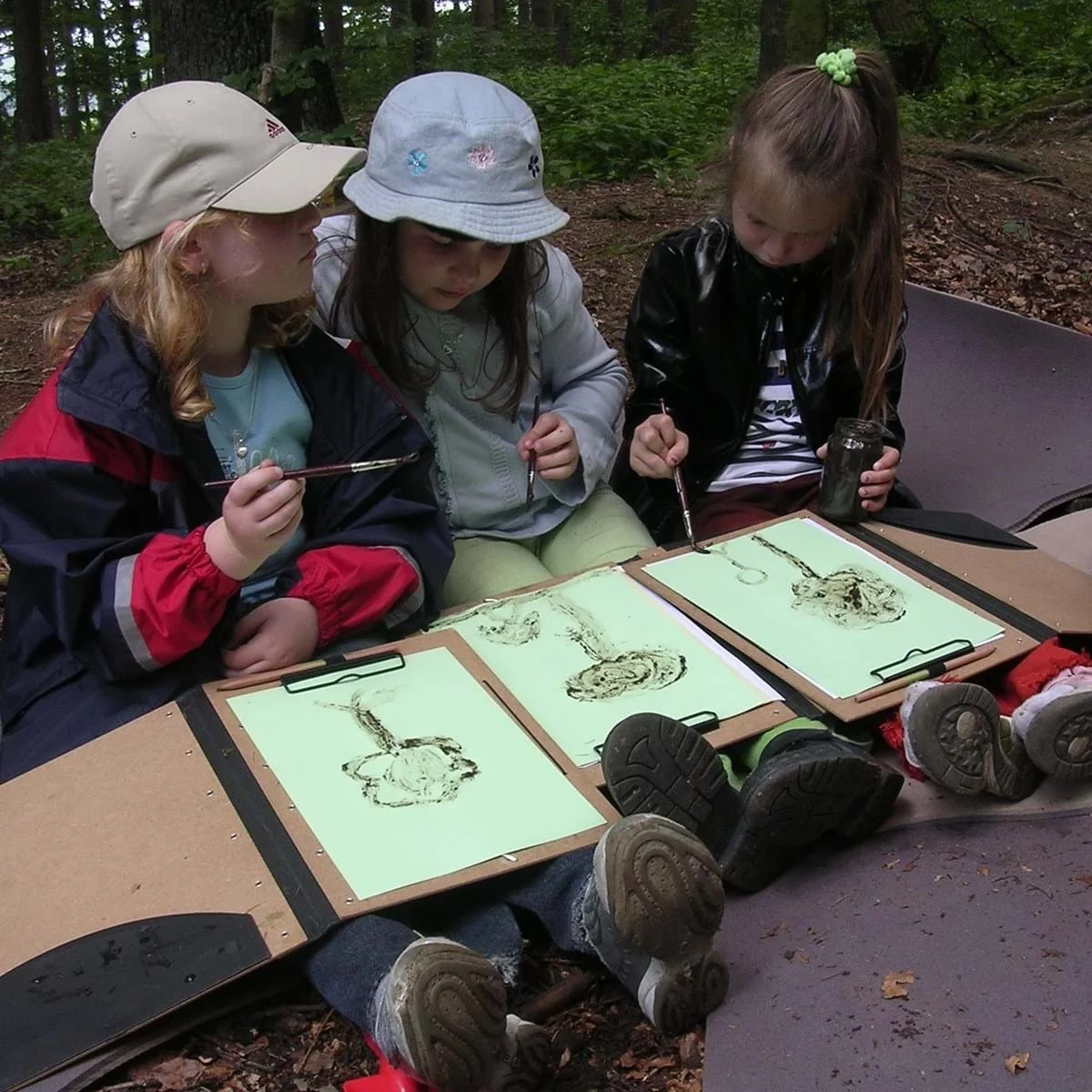 Drei Kinder sitzen im Wald auf einer Bank und zeichnen mit Stiften auf Klemmbrettern
